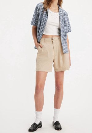 Femme portant un short beige, un crop top blanc, une chemise à manches courtes bleue à carreaux, des chaussettes blanches et des mocassins noirs, debout devant un fond uni.