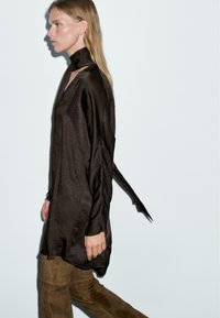 Tunika a maniche lunghe in satin marrone scuro con colletto alto e fessure laterali, abbinata a pantaloni in suede marrone, su uno sfondo chiaro.