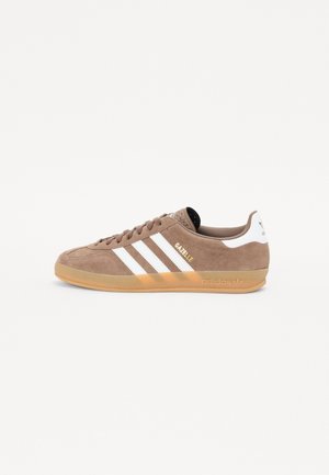 Baskets Adidas Gazelle marron avec des bandes blanches et une semelle en gomme, vue de côté sur un fond clair uni.