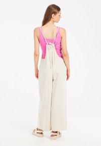Overoles beige con piernas anchas, tirantes ajustables y una camiseta rosa debajo. La tela parece ligera y texturizada. Calzado informal.