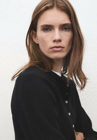 Cardigan nero con scollo rotondo e bottoni dorati, caratterizzato da una texture morbida e una vestibilità comoda. Il modello posa contro uno sfondo chiaro.