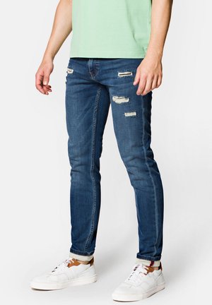 Slim-fit niebieskie dżinsy z przetarciami, z dziurami i postrzępionymi krawędziami. W połączeniu z białymi trampkami i zieloną koszulą.