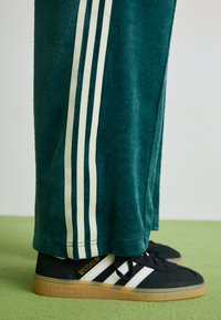 Green textured pants with white side stripes, paired with black Adidas sneakers featuring three white stripes and a gum sole.