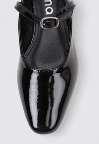 Zapato de charol negro con puntera redondeada, con una fina correa en el tobillo y un acabado brillante. El interior es de cuero negro mate.