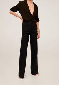 Kobieta w czarnej bluzce z głębokim dekoltem w serek i podwiniętymi rękawami, w wysokim stanie, w czarnych szerokich spodniach, stoi z jedną ręką na biodrze.