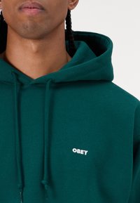 Zaļš kapuce ar mīkstu audumu, ar kangurupodu, regulējamu kapuci un baltu "OBEY" logo uz krūtīm.