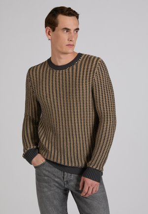 Homme portant un pull tricoté à motifs gris foncé et beige et un jean gris, regardant sur le côté avec une main dans la poche, sur un fond uni.