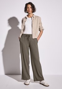 Beige knoopjack, witte T-shirt, olijfgroene wijdvallende broek en lichtgekleurde sneakers. Textuurstof en relaxte pasvorm.