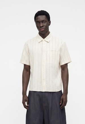 Jeune homme portant une chemise à manches courtes, de couleur claire, rayée verticalement et boutonnée, et un pantalon foncé et ample sur un fond uni.