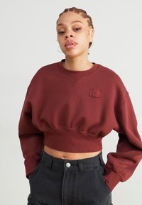 Burgundy színű cropped pulóver, kerek nyakkivágással, hajtott ujjakkal és egy kis logóval az elején. Magas derekú fekete nadrággal párosítva.