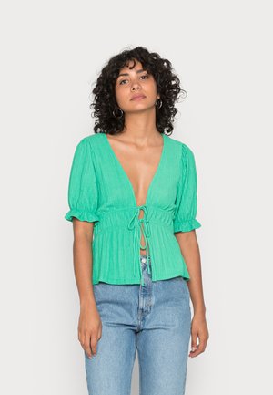 Blusa verde texturizada com decote em V profundo, mangas curtas bufantes e um laço na frente. Combinada com jeans de cintura alta em azul claro.