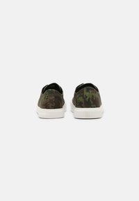 Une paire de baskets basses avec un motif de camouflage vert et marron et des semelles en caoutchouc blanches, vues de l'arrière sur un fond blanc.