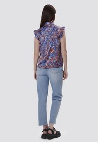 Korte mouw top met een blauw en oranje geometrisch patroon, met gerimpelde schouders; gecombineerd met lichtblauwe jeans en zwarte plateausandalen.
