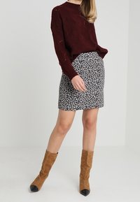 Suéter de punto burdeos, falda de leopardo gris, botines de ante marrón con puntera negra. La modelo está de pie frente a un fondo neutro.