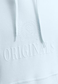 Sweat à capuche bleu clair avec texte embossé « ORIGINALS » et un emblème circulaire au-dessus avec des feuilles décoratives et un petit texte.