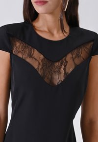 Donna che indossa un abito nero con maniche corte e un pannello in pizzo a forma di V sul petto, su uno sfondo semplice.