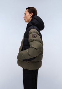 Puffjakke med sort hætte, olivengrøn krop og et rundt emblem på ærmet. Fremstillet af polstret materiale med en mat overflade.