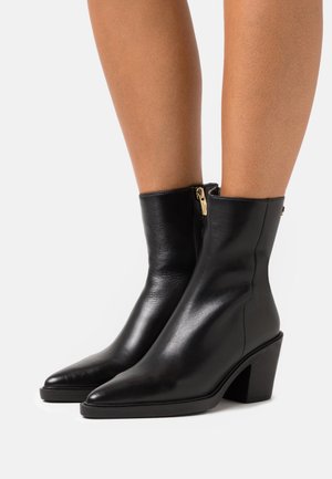Botas de cuero negro hasta el tobillo con puntas afiladas y tacones cuadrados. Cuentan con una cremallera lateral con herrajes dorados. Textura suave y diseño ajustado.