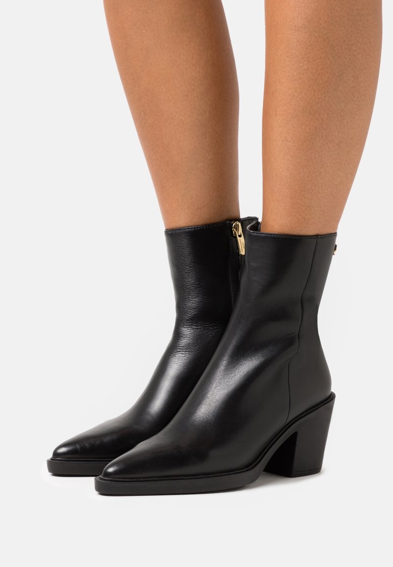 Botas de cuero negro hasta el tobillo con puntas afiladas y tacones cuadrados. Cuentan con una cremallera lateral con herrajes dorados. Textura suave y diseño ajustado.