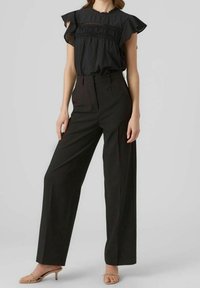 Sort top med flæser ved ærmerne og smock-detaljer, parret med højtaljede, brede sorte bukser og nude strophæle.