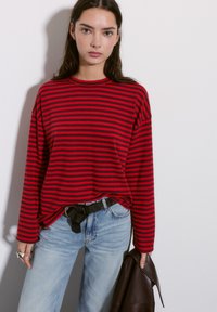 Red and navy striped long-sleeve shirt paired with light blue jeans, featuring a black belt and holding a dark brown jacket.