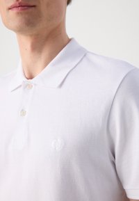 White cotton polo shirt featuring a classic collar, two-button placket, and subtle embroidered logo on the left chest. Textured fabric.