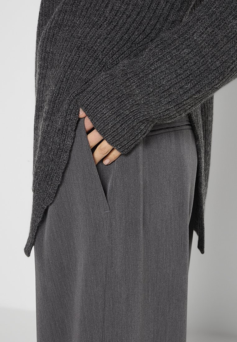 Maglione grigio a costine con una vestibilità ampia e uno spacco laterale, abbinato a pantaloni grigi su misura con una tasca discreta.