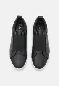 Czarne buty wsuwane z elastycznymi paskami, błyszczące skórzane wykończenie, okrągły czubek, biała gumowa podeszwa oraz logo "Calvin Klein" z boku.