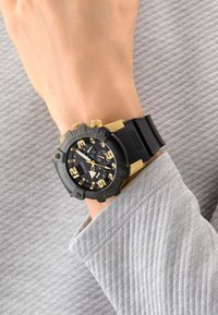 Orologio analogico nero e oro con cinturino in silicone, numeri grandi e caratteristiche cronografiche, mostrato su un polso, abbinato a una manica grigia con texture.