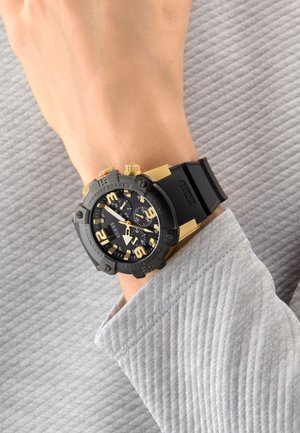Montre analogique noire et dorée avec un bracelet en silicone, grands chiffres et fonctionnalités chronographiques affichées sur un poignet, assortie à une manche grise texturée.