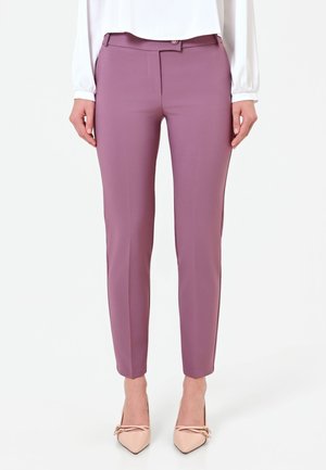 Donna con pantaloni aderenti color malva, camicetta bianca e tacchi beige con punta a contrasto, in piedi davanti a uno sfondo bianco semplice.