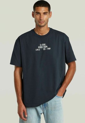 Homme portant un t-shirt noir avec un texte blanc et un jean bleu clair, debout avec une main dans la poche devant un fond uni.