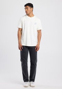 Witte katoenen T-shirt met korte mouwen en een blauwe bloemenafbeelding, gecombineerd met donkergrijze broeken en witte sneakers, geplaatst tegen een neutrale achtergrond.