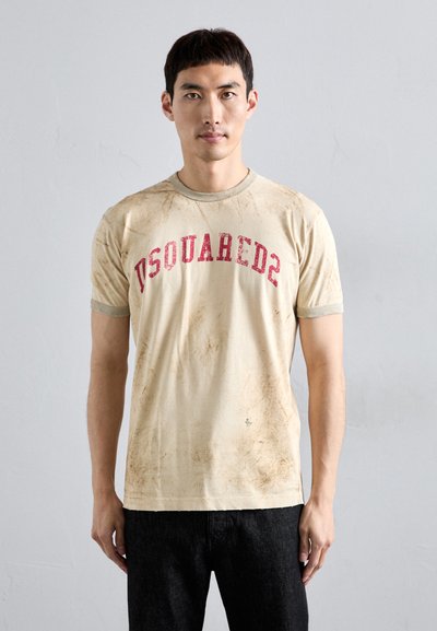 Beige Baumwoll-T-Shirt mit rotem "DSQUARED2"-Druck, kurzen Ärmeln und einem abgenutzten Textur. Kombiniert mit dunklen Jeans.