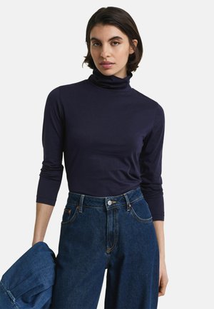 Vrouw in een marineblauwe turtleneck met driekwart mouwen en hoge taille donkere blauwe jeans, die in één hand een spijkerjack vasthoudt.