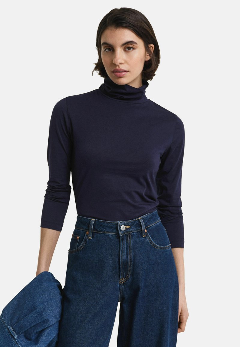 Vrouw in een marineblauwe turtleneck met driekwart mouwen en hoge taille donkere blauwe jeans, die in één hand een spijkerjack vasthoudt.