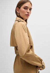 Beige Trenchcoat mit einem taillierten Bund, kurzen Kapuzenärmeln und Knopfakzenten. Glattes Material mit passgenauer Passform und minimalistischem Design.