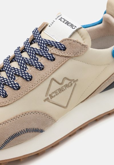 Beżowe trampki z granatowo-białymi wzorzystymi sznurówkami, wykonane z zamszu i materiałów tekstylnych, z wytłoczonym logo i teksturowaną gumową podeszwą.