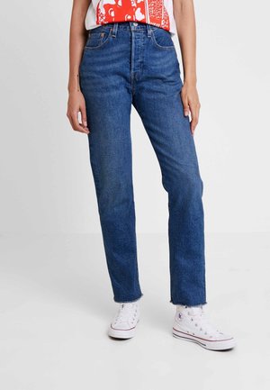 Personne portant un jean bleu taille haute avec des ourlets effilochés et des baskets montantes blanches, se tenant contre un fond clair uni.