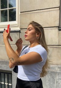 En kvinna med långt, vågigt blont hår bär en vit ribbad croptop och applicerar rosa läppglans med en röd handspegel.