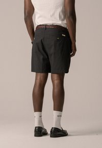 Pantaloni corti neri con vestibilità dritta, in tessuto di cotone, due tasche posteriori e una cintura intrecciata marrone. Indossati con scarpe nere e calze bianche alla caviglia.