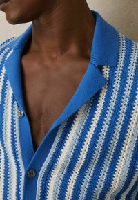 Blau-weiß gestreifter Strickcardigan mit strukturierter Oberfläche, ausgestattet mit einem blauen Kragen und Knöpfen, wobei eine Nahaufnahme des Stoffes hervorgehoben wird.
