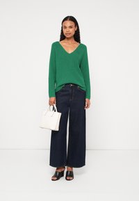 Žalias šifruotas megztinis V formos iškirpte, derinamas su plačiomis tamsiomis džinsomis, juodais atvirais bateliais ir mažąja balta rankine.