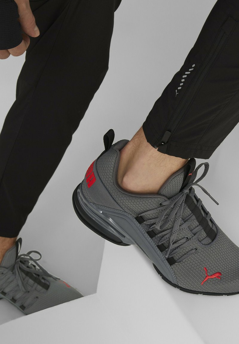 Chaussure de sport grise avec un dessus en mesh, accents noirs et logo rouge. Dotée d'une semelle rembourrée et d'un design à lacets. Portée avec un pantalon noir.