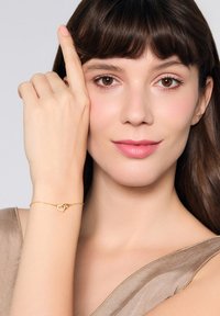 Złota bransoleta z sercowym wzorem, delikatnym łańcuszkiem i polerowanym wykończeniem, noszona na nadgarstku osoby na tle w neutralnych kolorach.