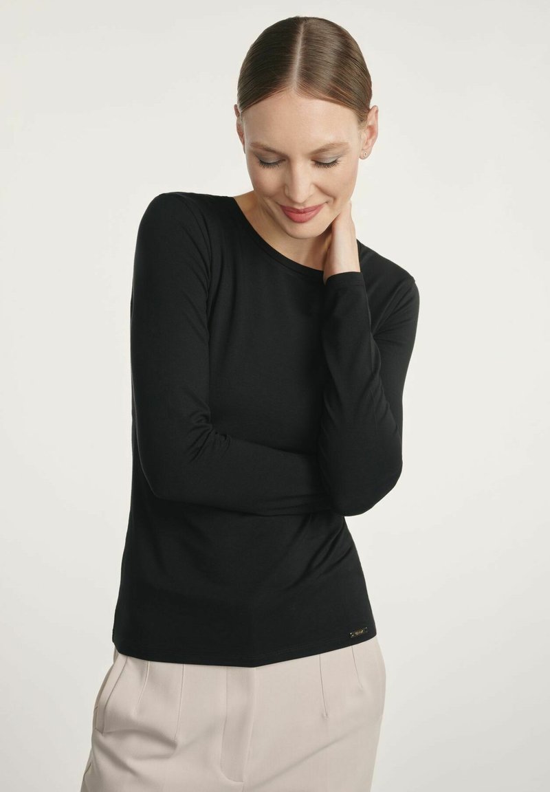 Czarny długi rękaw top z miękkiego materiału, z okrągłym dekoltem i dopasowanym krojem. Posiada subtelny detal z logo na dolnym brzegu. W połączeniu z jasnymi spodniami.