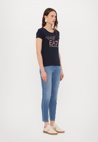 Tamsiai mėlyna trumpas rankovėmis marškinėliai su spalvinga "EAT EMPORIO ARMANI" grafika, derinami su šviesiai mėlynais aptemptais džinsais ir smėlio spalvos sportbačiais.