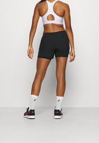 Svarta atletiska shorts med lös passform, kombinerade med en ljusrosa sport-BH med nyckelhålsdesign i ryggen. Vita och svarta sneakers med matchande strumpor.