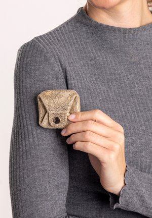 Hand hält kleinen, strukturierten Goldbeutel mit Knopfverschluss vor grauem, geripptem Langarmshirt.