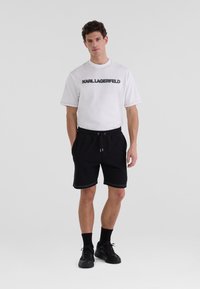 Vit t-shirt med svart "KARL LAGERFELD"-logotyp, i kombination med svarta shorts. Outfitten har en avslappnad passform och kontrasterande sömmar.